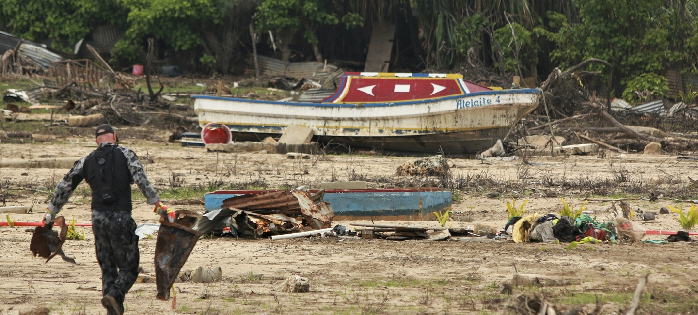 a person cleans up debris on the island of Tonga after a volcanic eruption in 2022 caused a tsunami. A boat is on land.