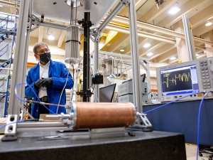 A faculty member wearing a face mask and a blue lab coat stands in a laboratory. He stands behind a copper tube, some silver scaffolding, and a flashing screen.
